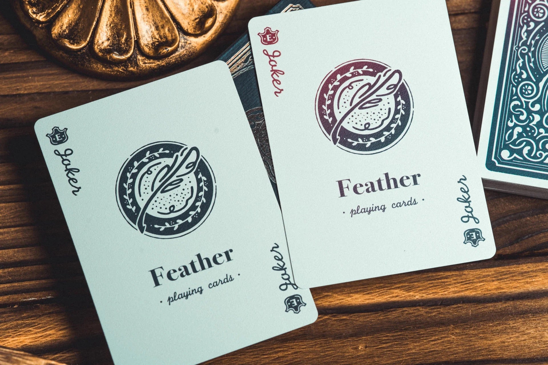 Two Feather playing cards laid on a wooden surface, with part of a teal patterned card deck visible at the right edge and a decorative bronze object at the top left.