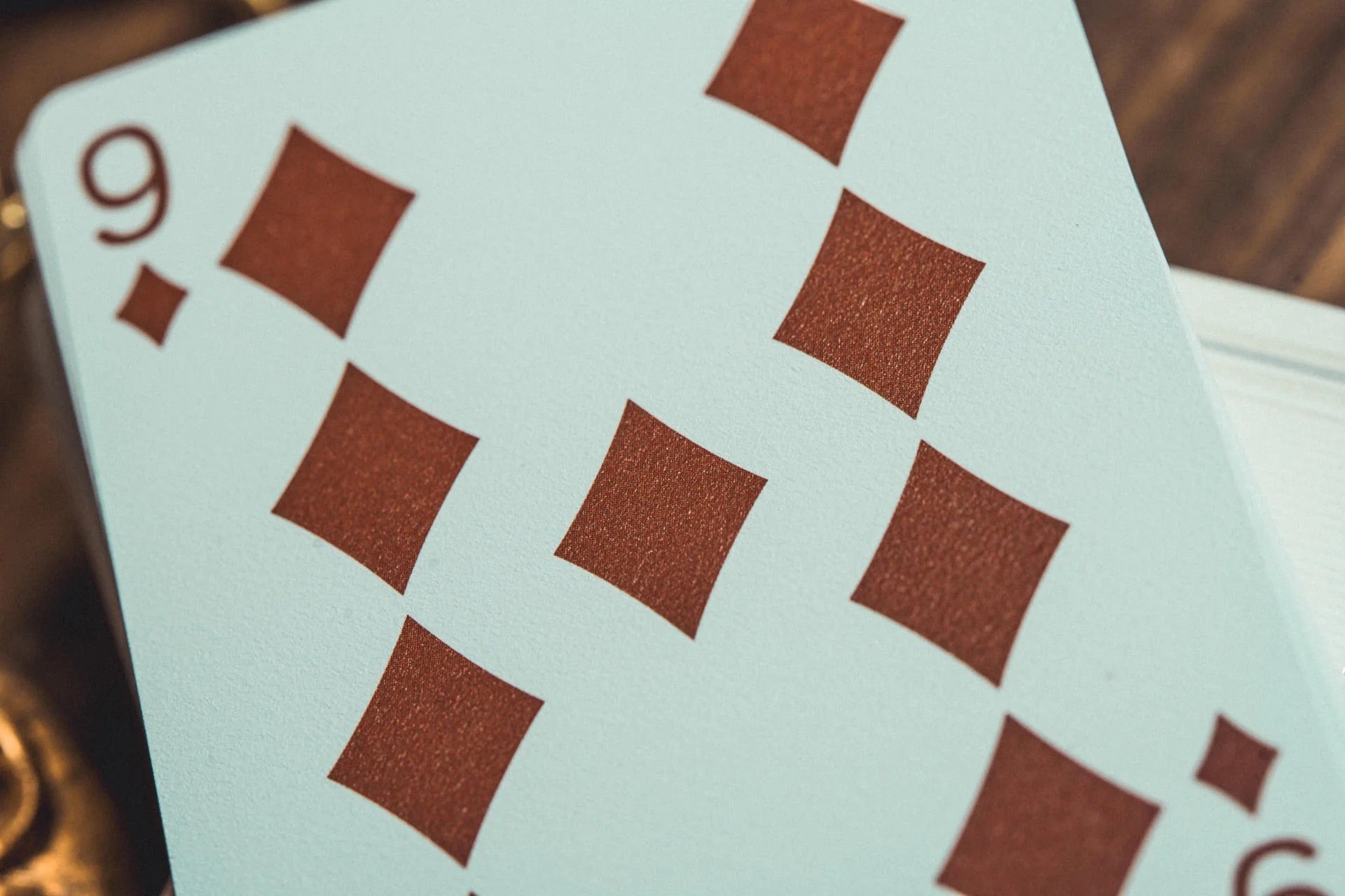 A close-up shot of a 9 of diamonds playing card, showing the light blue card face with brown diamond suits, resting on a dark wooden surface.