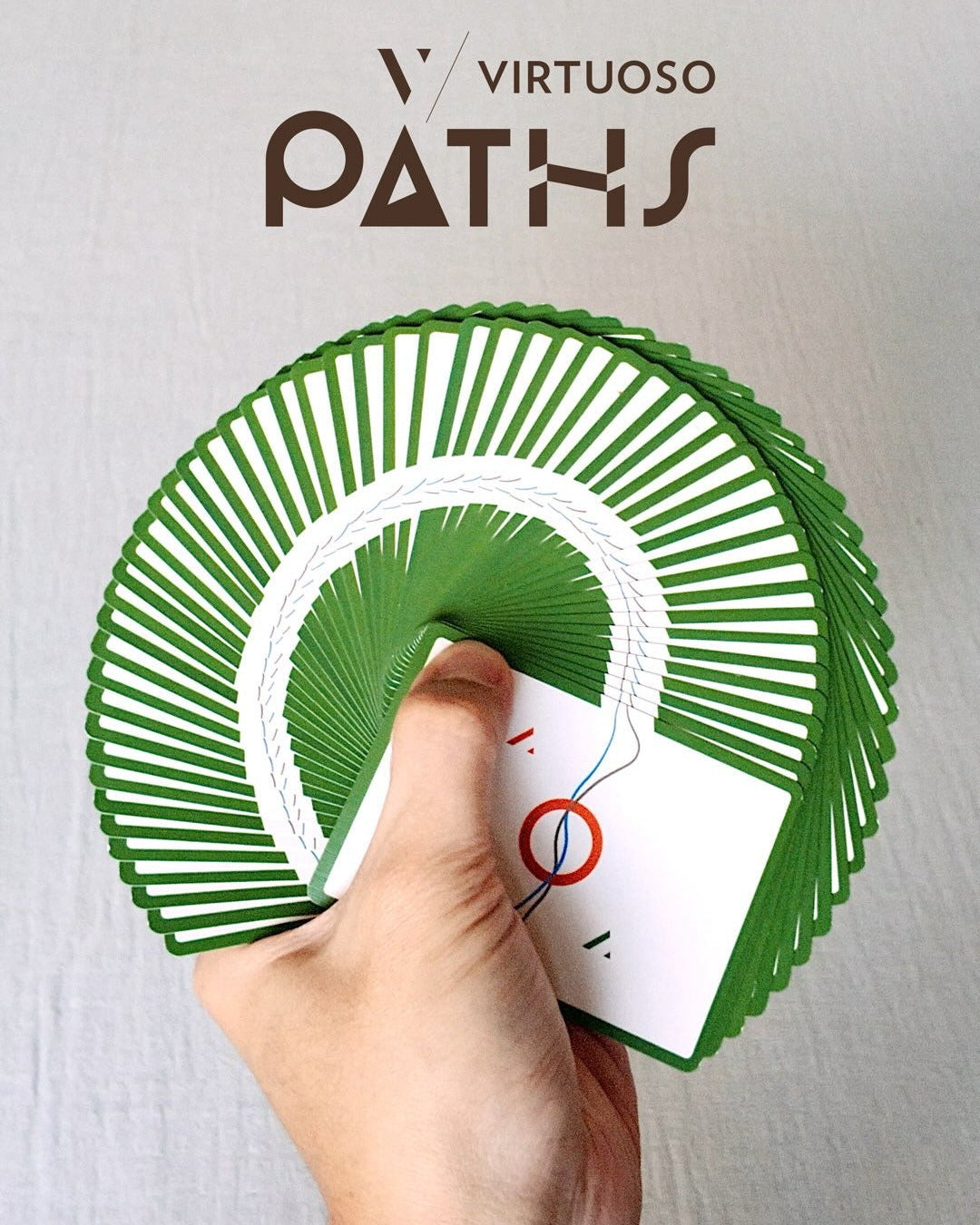 A hand holds a fanned deck of cards from the brand Virtuoso Paths, showing a green and white design. The brand logo &