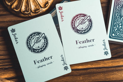 Two Feather playing cards laid on a wooden surface, with part of a teal patterned card deck visible at the right edge and a decorative bronze object at the top left.