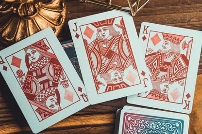 Three red-backed playing cards (Queen, Jack, and King) are arranged on a wooden surface next to a stack of cards showing blue and red backs, with a decorative gold object in the background.
