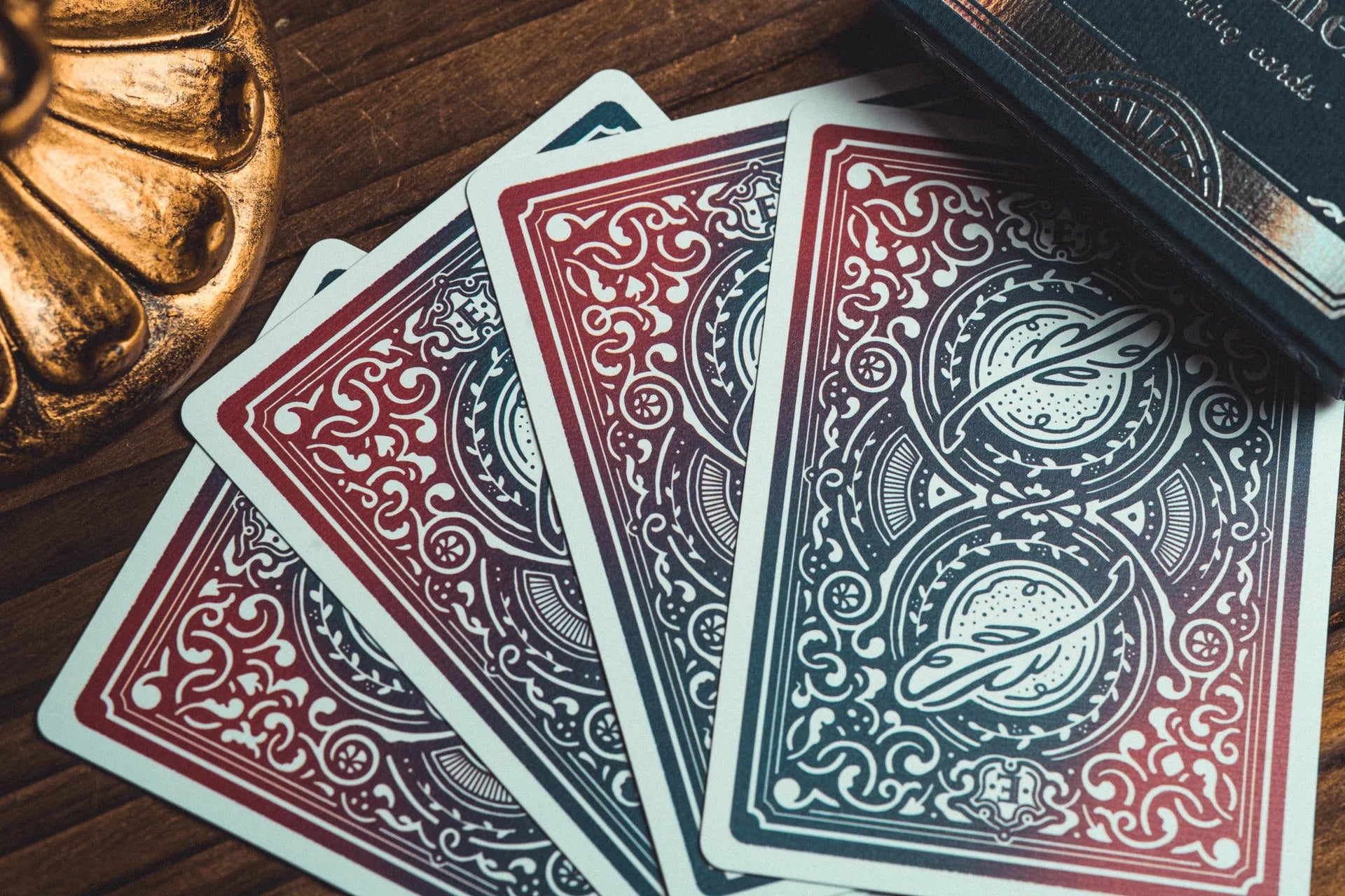 Four fanned-out playing cards with ornate red, blue, and white decorative back designs, arranged on a dark wooden surface next to a gold-toned decorative object and a dark book or box.
