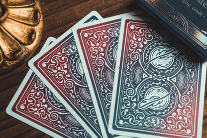 Four fanned-out playing cards with ornate red, blue, and white decorative back designs, arranged on a dark wooden surface next to a gold-toned decorative object and a dark book or box.