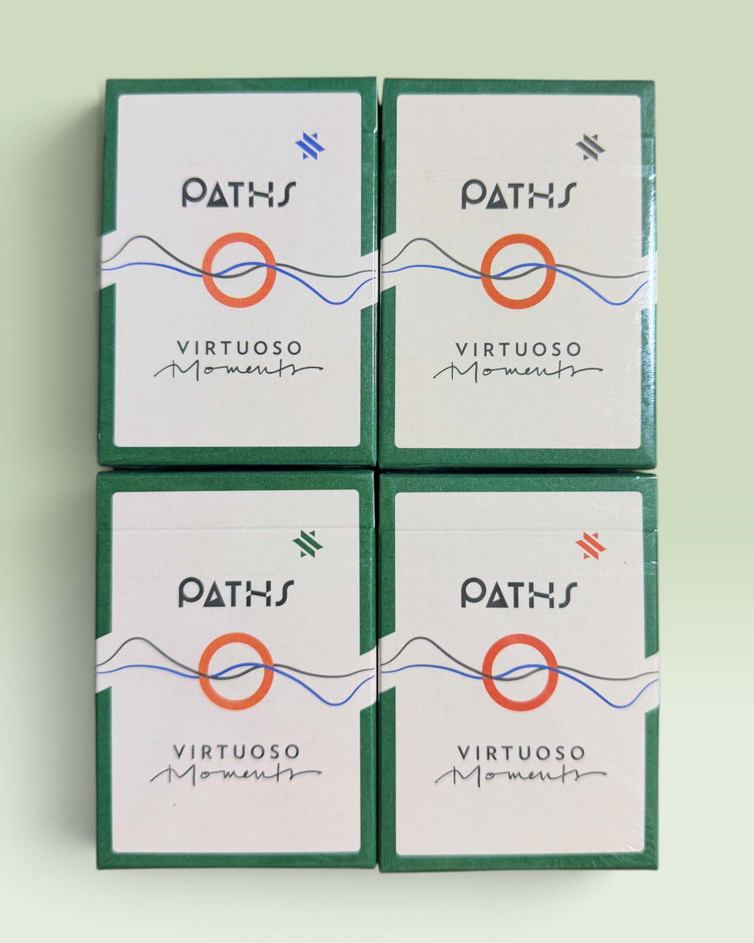 Four identical boxed cards arranged in a 2x2 grid on a pale mint background. The boxes are dark green with white front panels, featuring the brand name PATHS and the product name Virtuoso Moments, plus a central orange circle and blue line graphic.